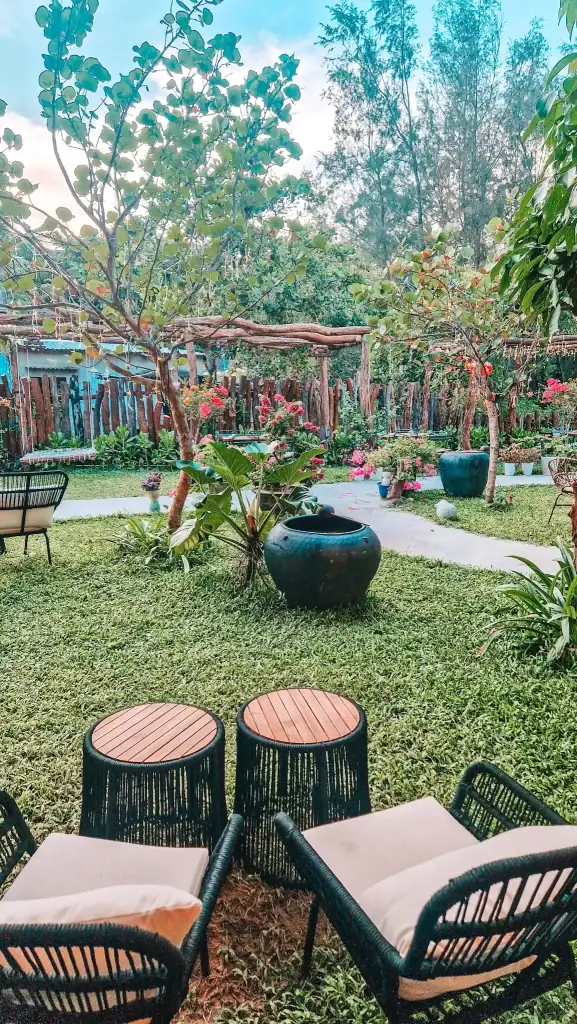 A cozy garden nook in An Bang with cushioned rattan chairs and wooden-topped tables, surrounded by potted plants, flowering shrubs, and trees for a peaceful cafe experience close to nature.