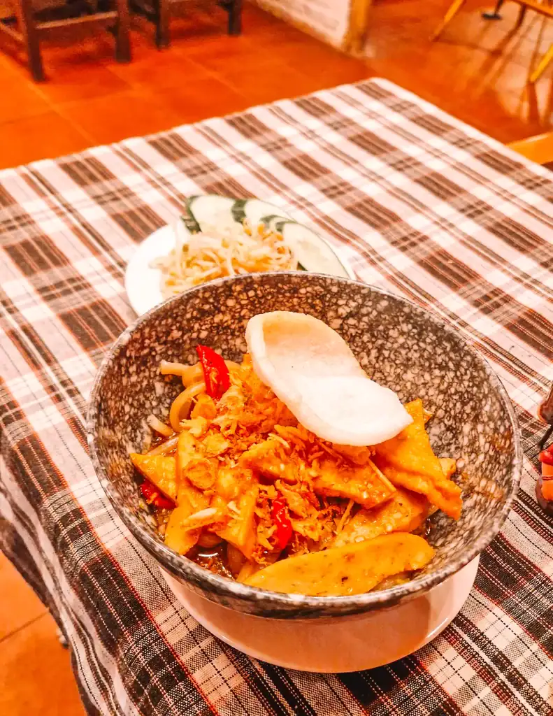 A bowl of stir-fried noodles with tofu, chili, and crispy toppings is served on a plaid tablecloth inside one of the cozy cafes in An Bang, with a side dish of fresh cucumber and bean sprouts.