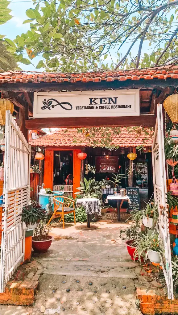 Entrance to Ken Vegetarian & Coffee Restaurant, one of the charming cafes in An Bang, with a tiled roof, white gate, and lush potted plants welcoming guests to its outdoor seating area.
