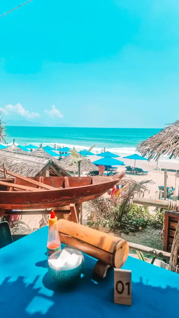 A beachfront cafe table in An Bang overlooks the sea, with blue umbrellas and thatched roofs dotting the sandy beach. A bowl of napkins, chili sauce, and table number '01' add to the casual charm of seaside cafes in An Bang.