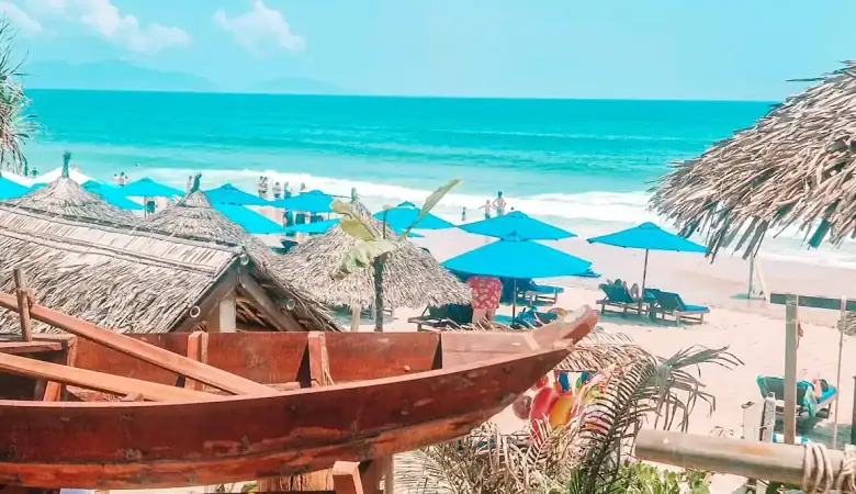 A beachfront cafe table in An Bang overlooks the sea, with blue umbrellas and thatched roofs dotting the sandy beach. A bowl of napkins, chili sauce, and table number '01' add to the casual charm of seaside cafes in An Bang.