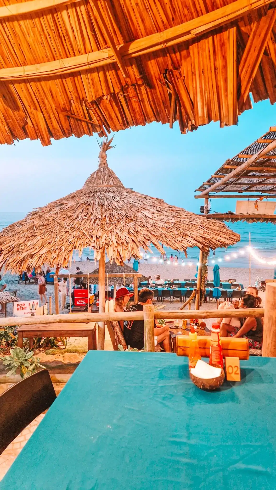 A beachfront table at one of the cafes in An Bang with a thatched umbrella roof, overlooking a lively beach scene at sunset. Bottles of chili sauce and napkins sit on a teal tablecloth as guests relax near the ocean under soft string lights.
