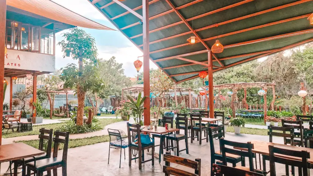 Open-air seating at one of the charming cafes in An Bang, featuring wooden tables under a canopy with garden views, colorful lanterns, and lush tropical greenery creating a serene ambiance.