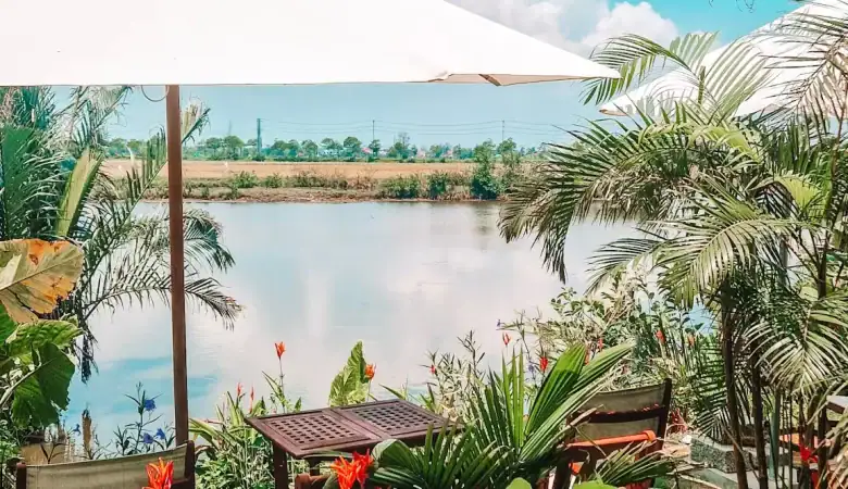 A cozy outdoor café by a calm river features wooden tables shaded by large white umbrellas, surrounded by tropical plants and bright red flowers, all under a clear blue sky.