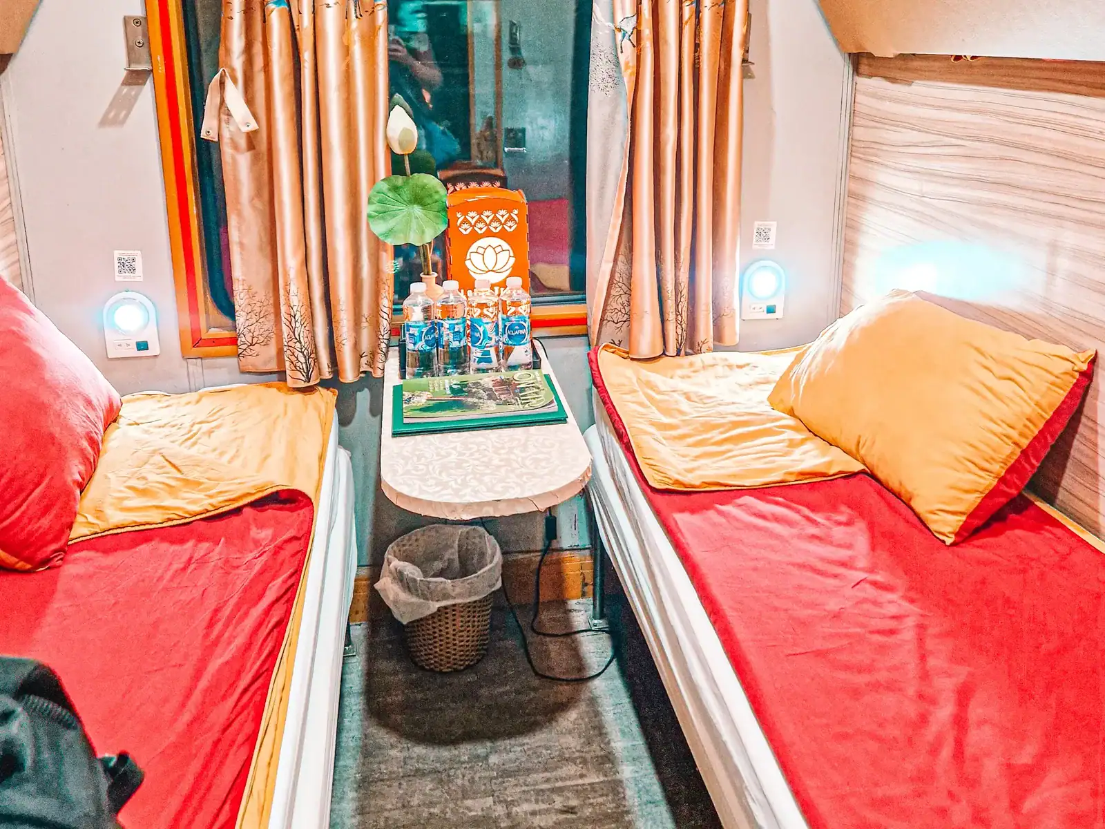 A cozy train cabin with two parallel beds covered in red and yellow bedding, a small table in between with bottled water and a lotus flower, and soft lighting illuminating the space.