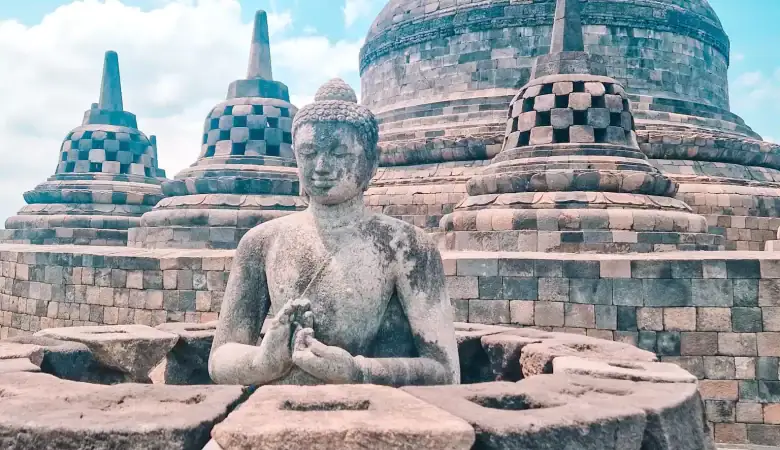 A serene stone Buddha statue seated in a lotus position at the top of Borobudur temple, surrounded by bell-shaped stupas under a bright blue sky.