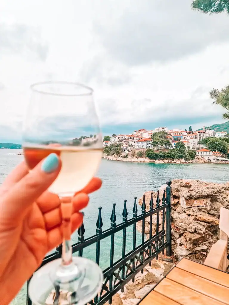 A hand holds a glass of white wine against a backdrop of charming coastal houses and calm blue waters in Skiathos, blending relaxation with picturesque town views.