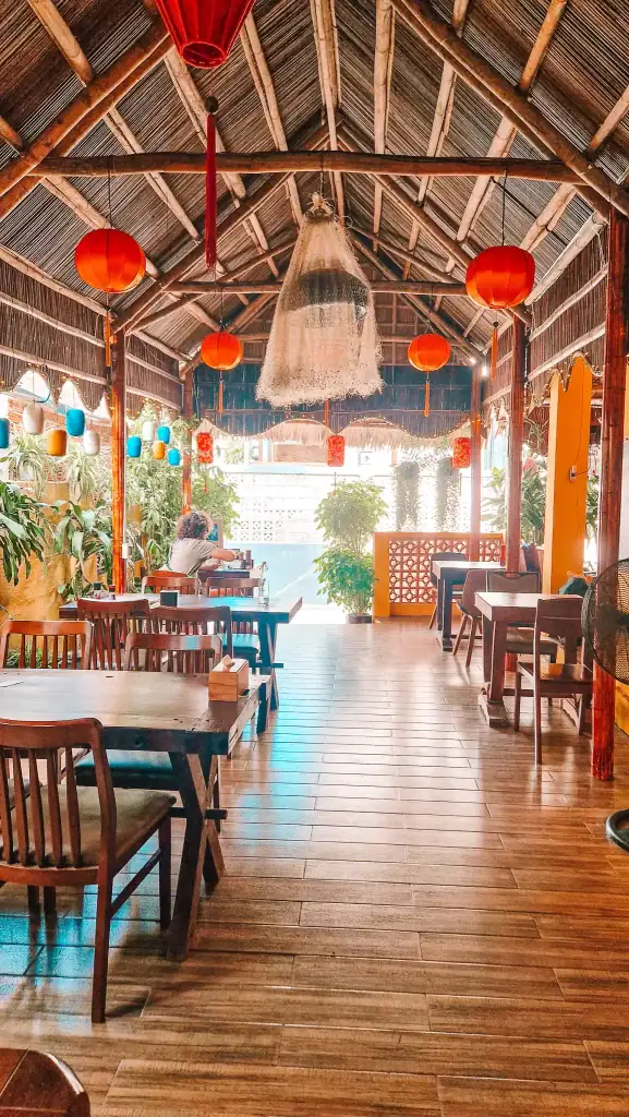 Interior of a bamboo-roofed cafe in An Bang, with wooden furniture, hanging lanterns, and greenery, offering a peaceful and stylish space for guests to relax and enjoy drinks or meals.