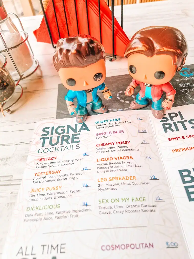 Two pop culture figurines are posed above a cheeky cocktail menu featuring bold drink names like “Liquid Viagra” at Crazy Rooster bar in Skiathos.