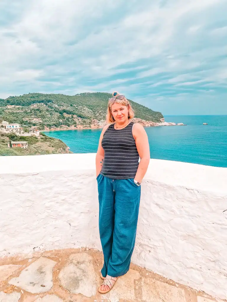 Em stands confidently in front of a whitewashed wall, with the vivid turquoise sea and Skopelos’ lush green hills stretching behind her under a partly cloudy sky.