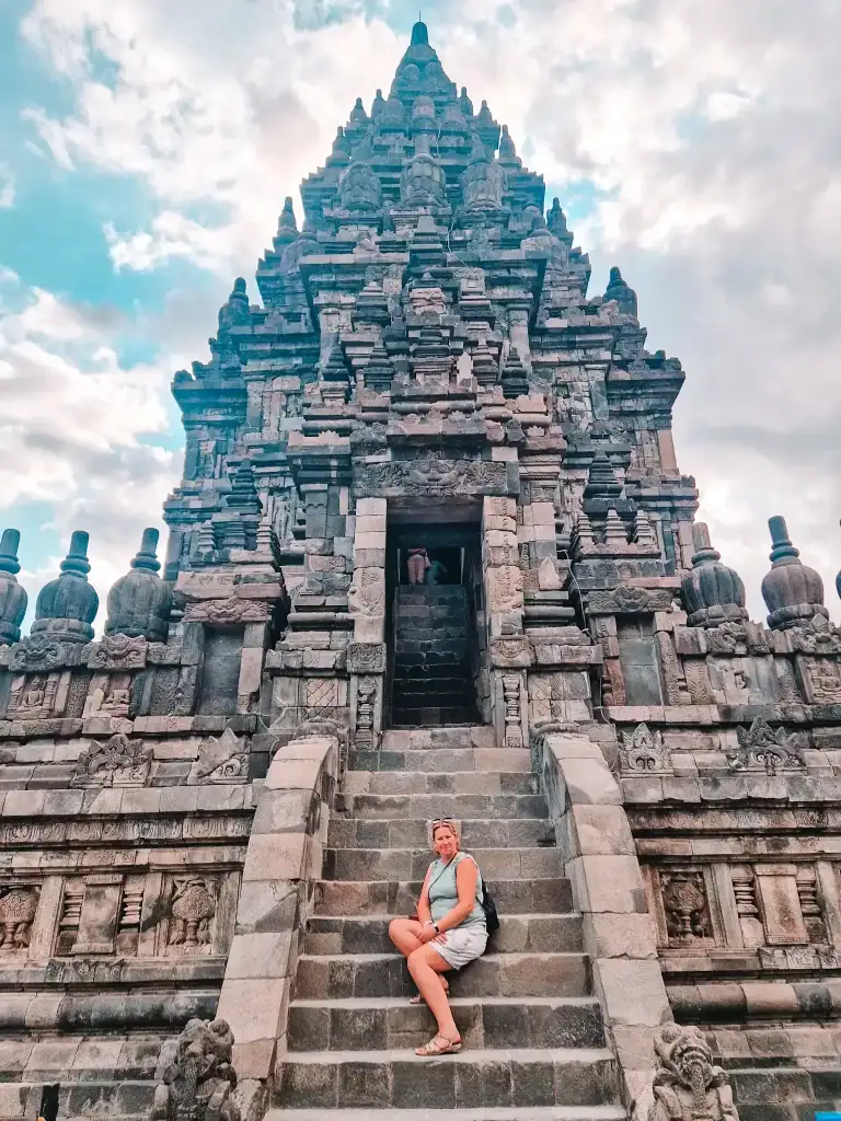 A woman sits on the stone steps of Prambanan Temple near Yogyakarta, Indonesia, framed by the temple's towering, intricately carved stone structure under a partly cloudy sky. This iconic Hindu temple is a must-see stop on a 2 days in Yogyakarta itinerary, showcasing ancient architecture and cultural heritage.