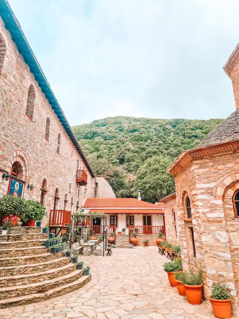 The peaceful stone courtyard of Evangelistria Monastery in Skiathos features terracotta planters, colorful flowers, and a backdrop of forested hills—offering a serene escape and one of the more spiritual things to do in Skiathos.