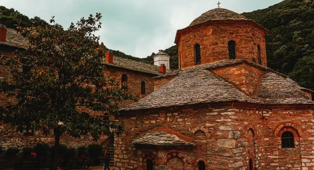 The historic Evangelistria Monastery in Skiathos stands with its stone walls, arched windows, and slate roofs set against a wooded mountain backdrop, offering a serene cultural stop.