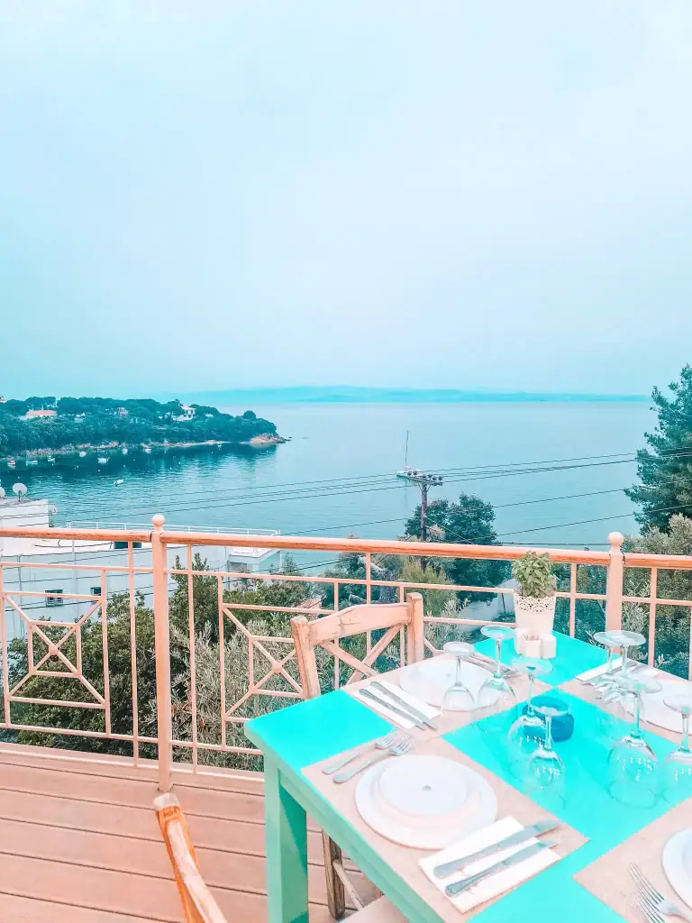 A turquoise table is elegantly set for four on a terrace at Infinity Blue restaurant, with panoramic views of the calm Aegean Sea and anchored boats below.