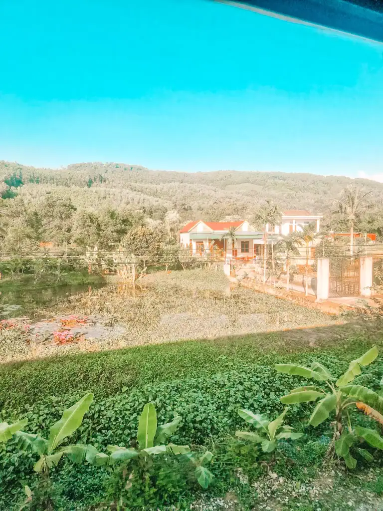 A scenic view from the train window, showing lush green fields, banana plants, and a large countryside house with a red roof. Rolling hills and a bright blue sky stretch across the background.