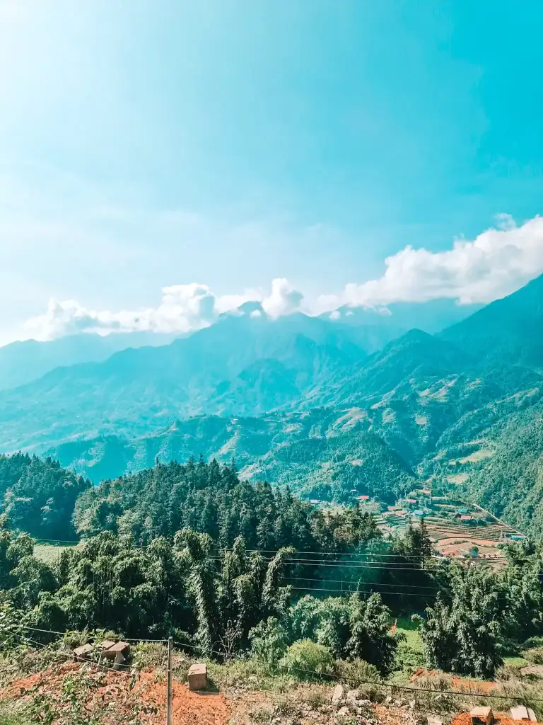A scenic view of rolling green mountains partially covered in mist, with scattered houses and terraced fields nestled in the valley. The bright blue sky and fluffy white clouds add to the serene atmosphere.