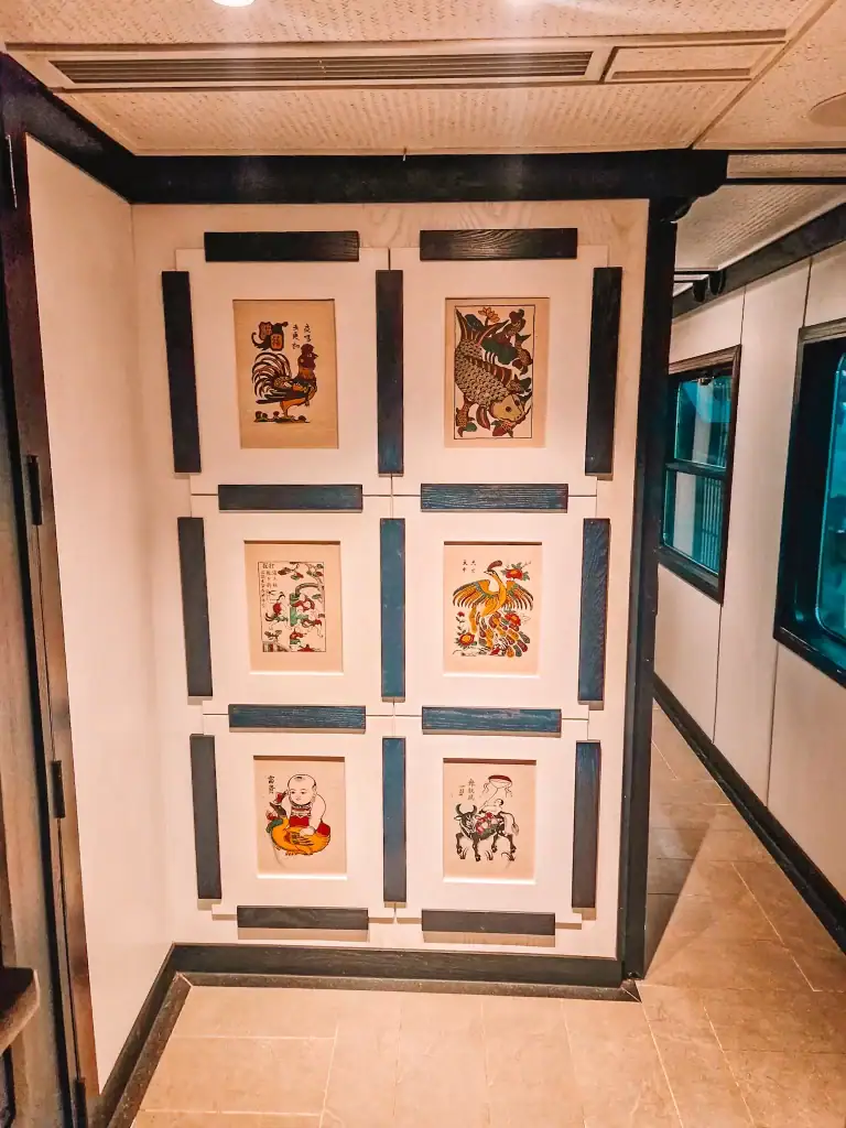 A section of a train's interior showcasing six framed traditional Vietnamese Đông Hồ paintings mounted on a beige wall with dark wooden borders. The paintings depict animals, folklore characters, and intricate designs.
