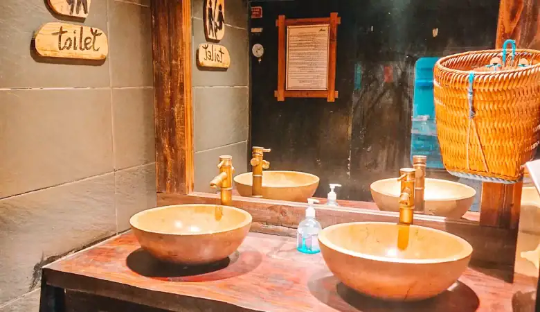 A rustic-style train washroom with a wooden vanity, two round stone sinks, and bronze faucets. Above the sink, a large mirror reflects the space, and wooden signs reading "toilet" in English are mounted on the dark tiled wall. A wicker basket hangs on the side, and a bottle of hand soap is placed on the countertop.
