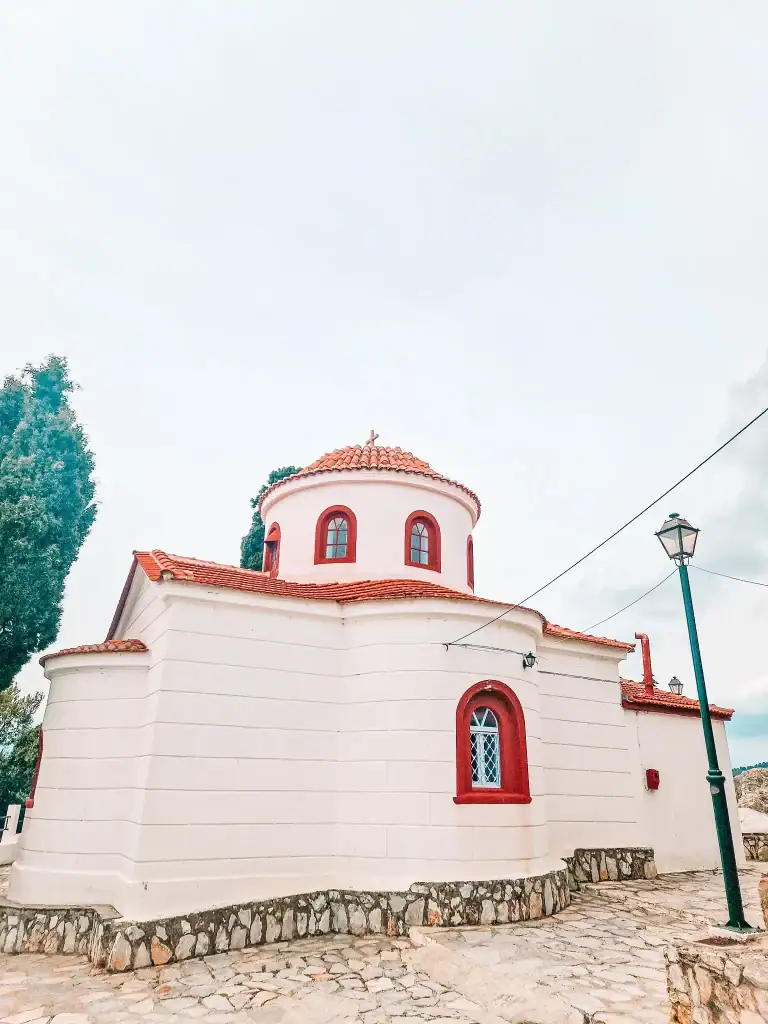 A small whitewashed church with red trim and a tiled dome sits on a stone-paved courtyard, its rounded architecture simple yet striking against the cloudy sky.