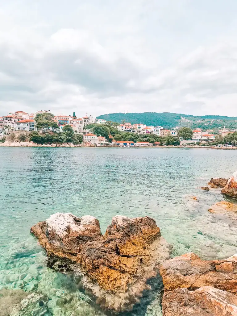 Golden rocks jut out from crystal-clear water in the foreground, while the pastel-colored buildings of Skiathos Town climb the shoreline across the bay.