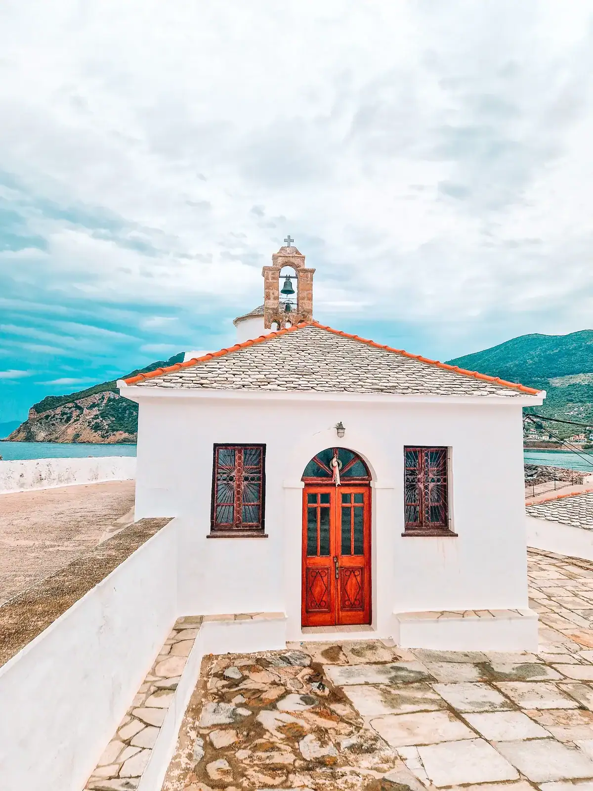 A quaint white Greek Orthodox church with a red door and bell tower sits near the sea, framed by mountain and ocean views, typical of Skopelos’ spiritual charm.