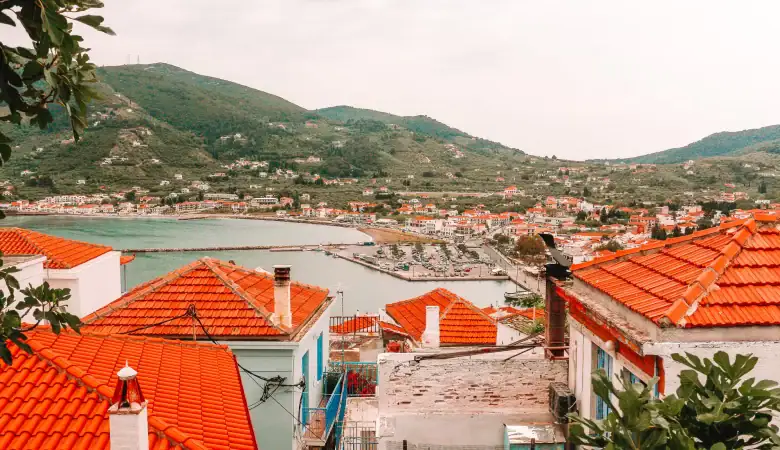 A sweeping view of Skopelos Town with terracotta rooftops and a small harbor nestled between green hills, capturing the laid-back beauty that draws visitors from neighboring Skiathos.