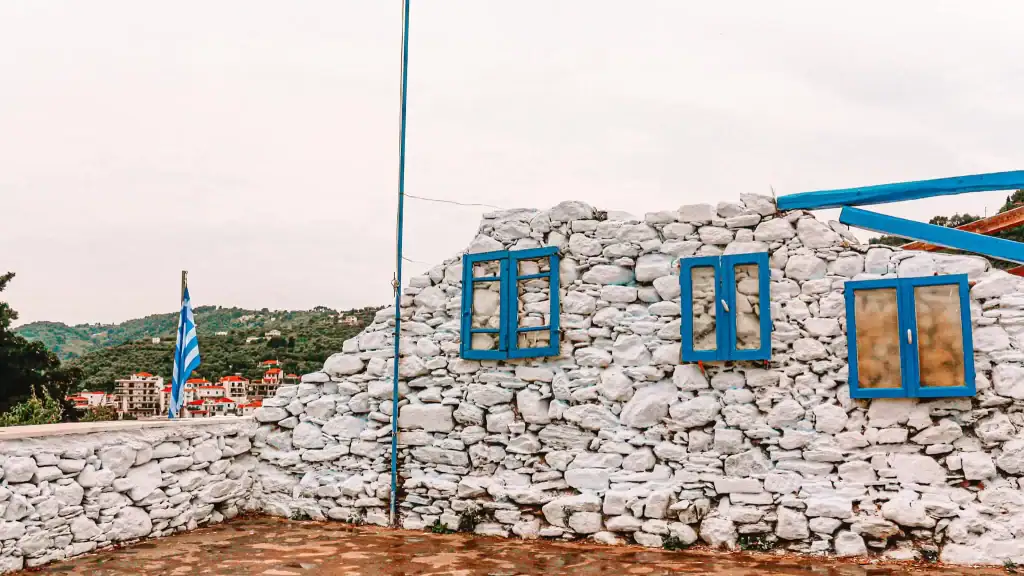 A rustic whitewashed stone wall with bright blue window shutters stands under a cloudy sky, with the Greek flag waving beside it and a hillside town visible in the background.