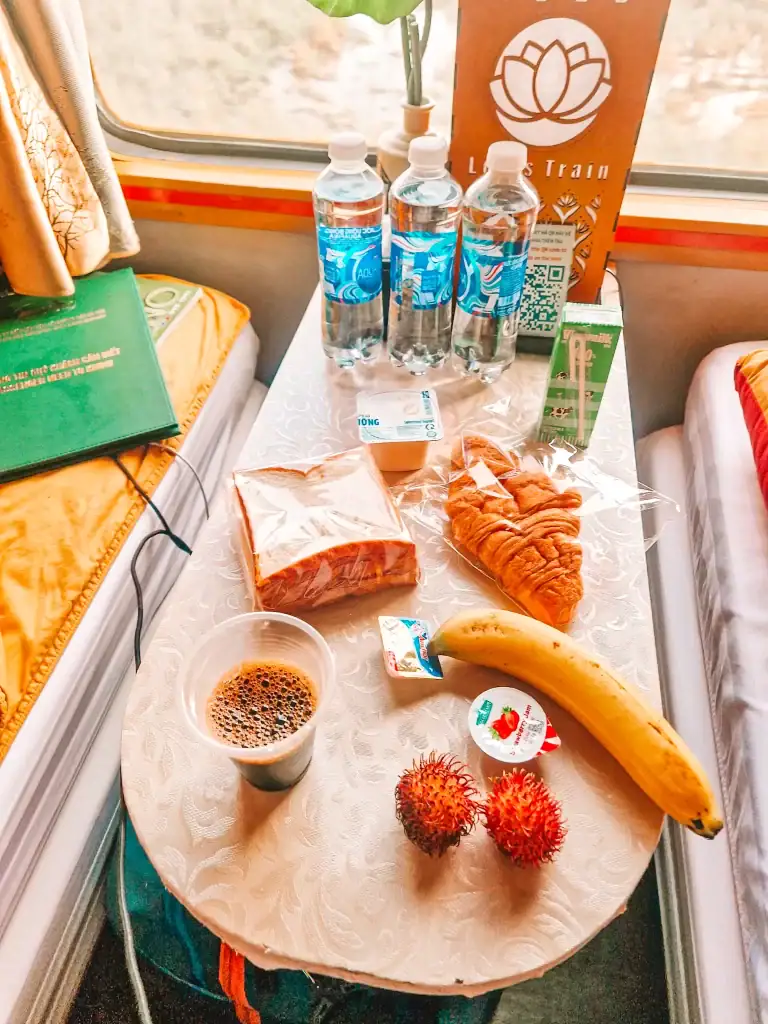 A small table inside a train cabin displaying a breakfast spread, including a cup of black coffee, a banana, two rambutan fruits, a croissant, a sandwich in plastic wrap, butter, jam, and yogurt. Three bottles of water and a green book rest nearby, with a decorative lotus-themed sign in the background.