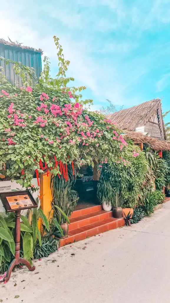 Entrance to Cosy Corner Café in An Bang, Vietnam, with a thatched roof, terracotta steps, and an overhanging canopy of pink bougainvillea and greenery framing the inviting cafe front.