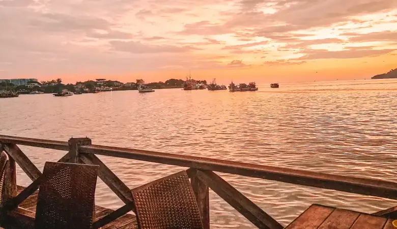 Waterfront dining setup with rattan chairs and candlelight on a wooden deck, overlooking a serene pink-and-orange sunset over the South China Sea — a romantic hidden gem in Kota Kinabalu for sunset lovers.