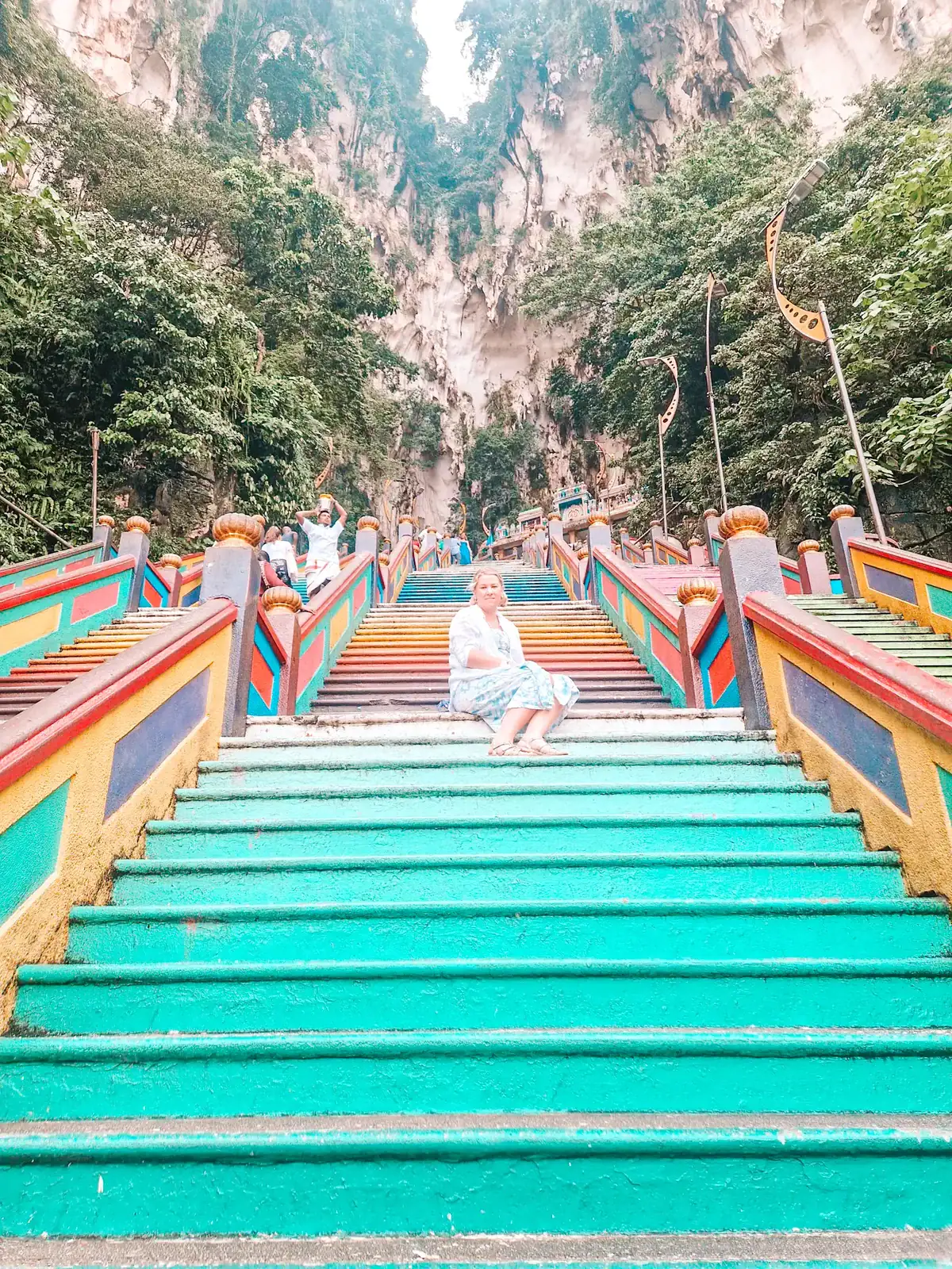 Em sits on the vibrant, rainbow-colored staircase of the Batu Caves, surrounded by towering limestone cliffs and tropical greenery—an iconic stop on any 3 week Malaysia itinerary.