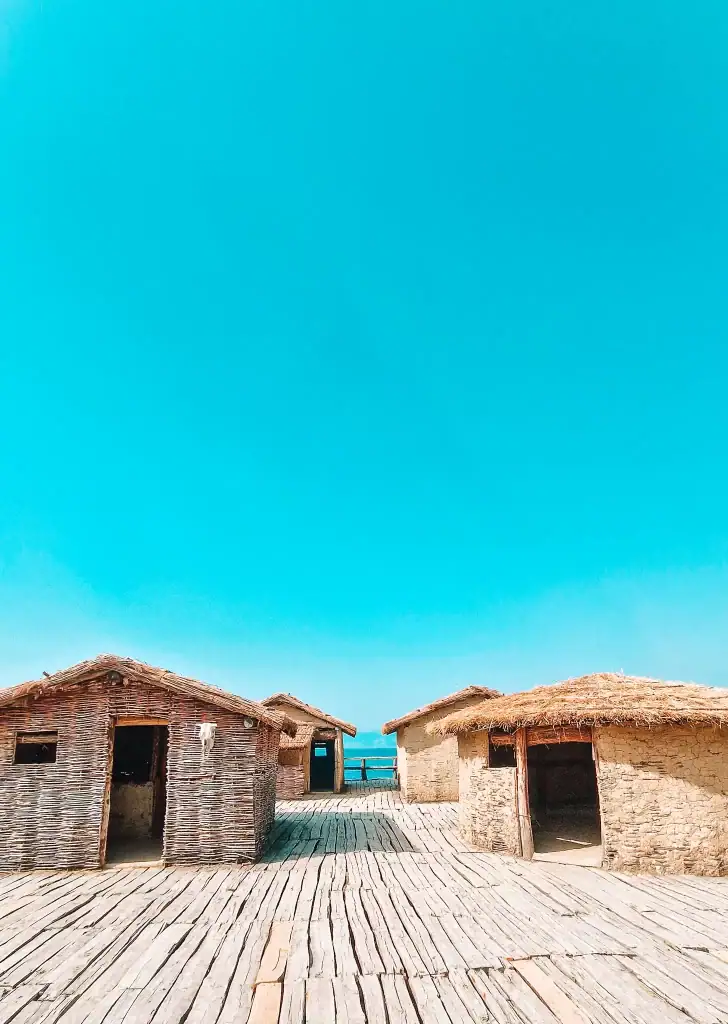 A group of reconstructed prehistoric huts with thatched and woven exteriors sit atop a wooden platform on Lake Ohrid, under a cloudless turquoise sky.