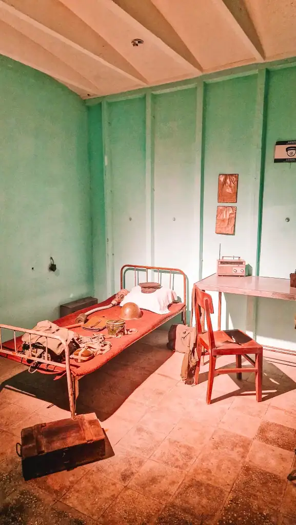 A re-creation of a Cold War-era officer’s room with a metal-framed bed, vintage desk and chair, a gas mask, helmet, and old radio—capturing life inside the Albanian bunker during a 2-day Tirana visit.