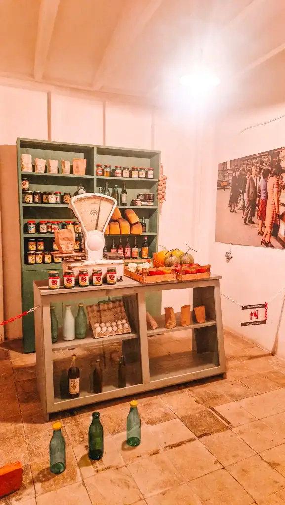 A preserved mock-up of a communist-era grocery shop featuring shelves of canned goods, eggs, loaves of bread, vintage glass bottles, and an old-fashioned scale.