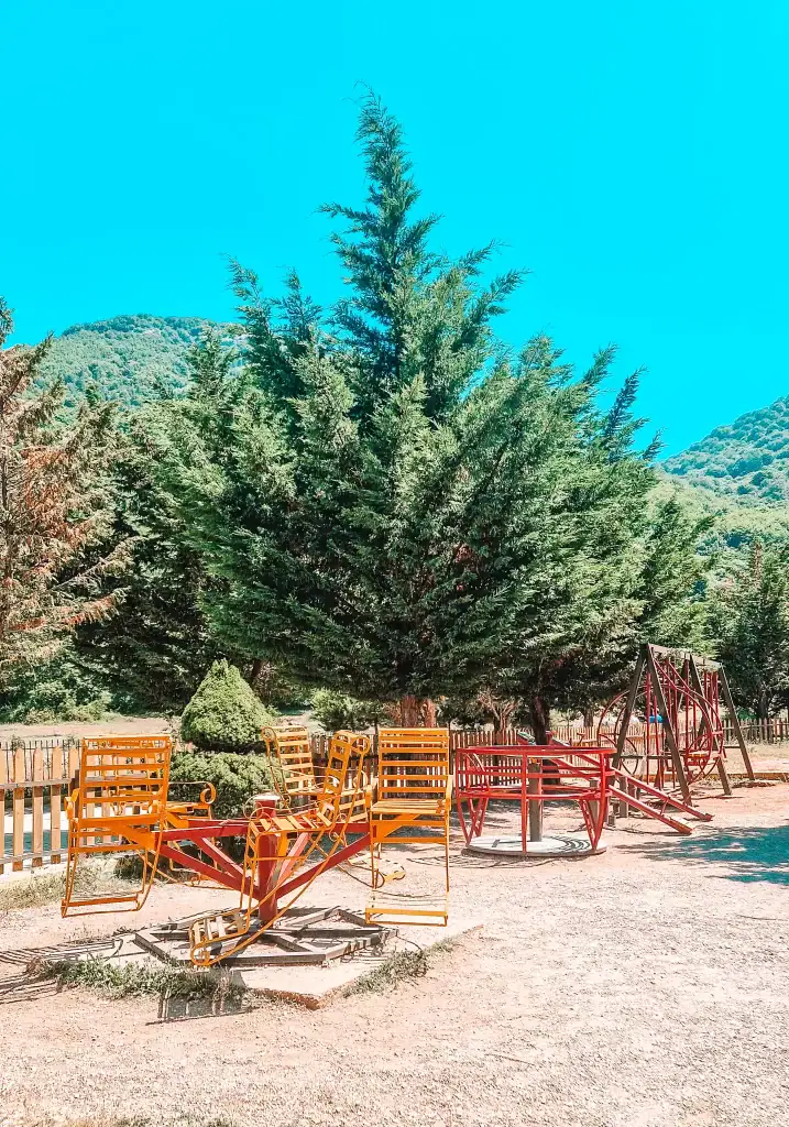 Colorful, slightly rustic playground equipment including merry-go-rounds and swings, surrounded by trees and mountain views near the Dajti cable car summit.
