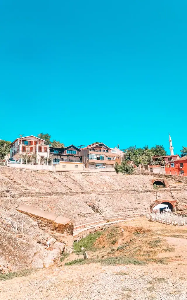 Ancient stone ruins of an amphitheatre sit at the base of a hill with a few colourful houses and a mosque visible above.
