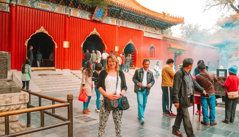 A woman smiles in front of the vibrant red entrance of the Lama Temple in Beijing, with other visitors walking around and incense burning nearby.