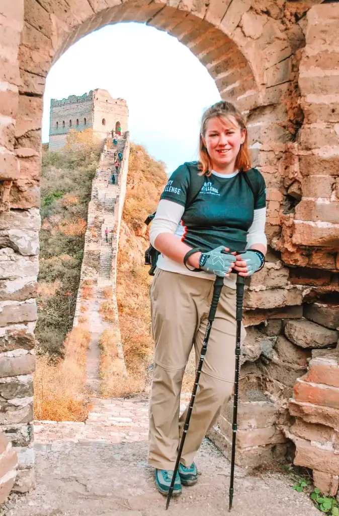 A woman in hiking gear rests inside a stone archway along the Great Wall of China, with steep steps and a watchtower behind her.