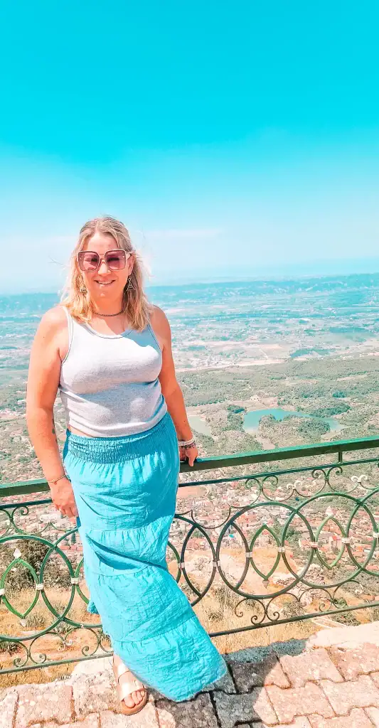 Em poses by a green railing at a high viewpoint near Kruje, with rolling hills, small lakes, and a patchwork of villages and farmland sprawling below.