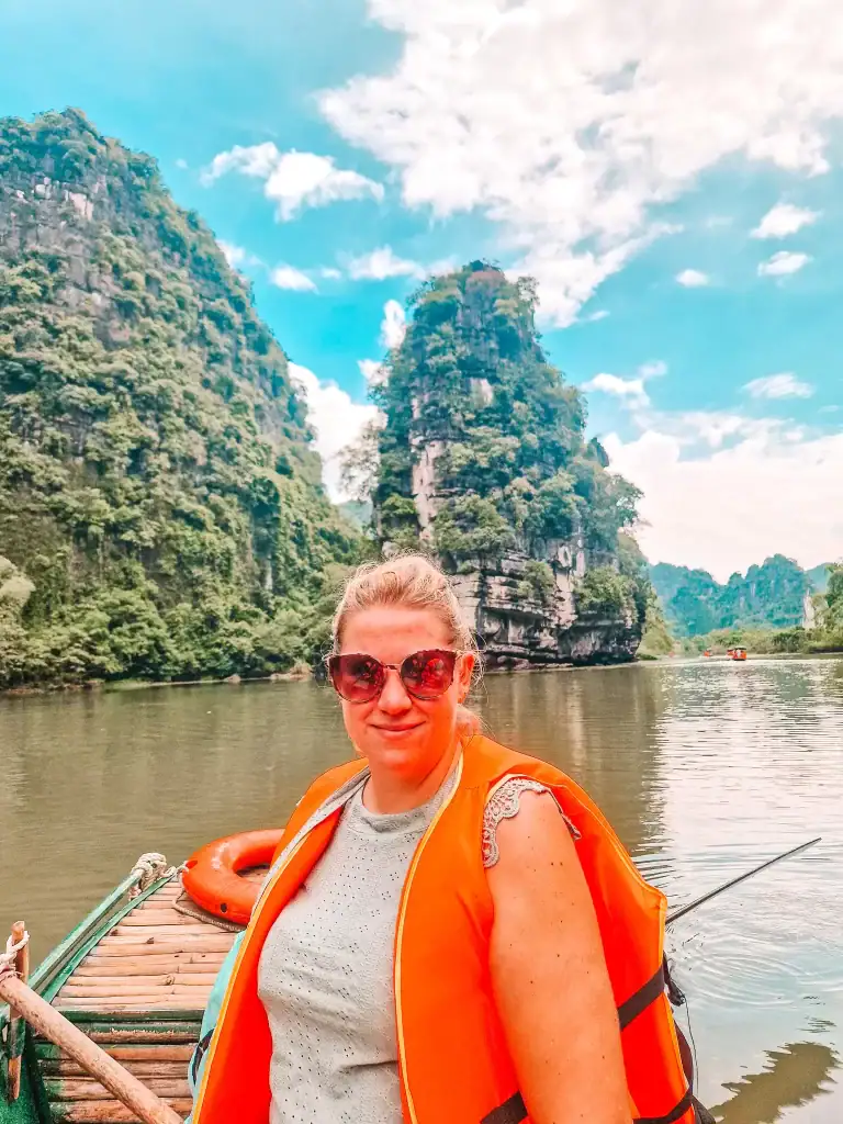 A woman wearing sunglasses and an orange life vest poses on a wooden boat in front of towering limestone karsts along the river in Ninh Binh, Vietnam. The dramatic cliffs and calm water highlight the region’s natural beauty.