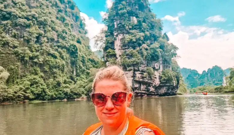 A woman wearing sunglasses and an orange life vest poses on a wooden boat in front of towering limestone karsts along the river in Ninh Binh, Vietnam. The dramatic cliffs and calm water highlight the region’s natural beauty.