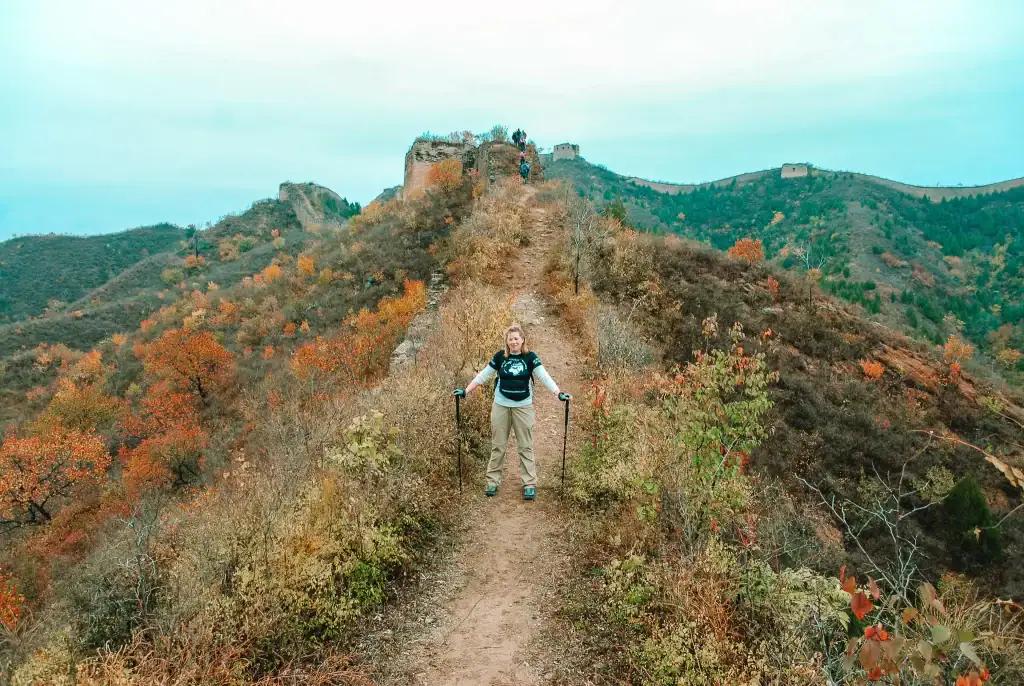 Em stands on a remote, rugged section of the Great Wall of China surrounded by autumn-colored hills and crumbling towers.