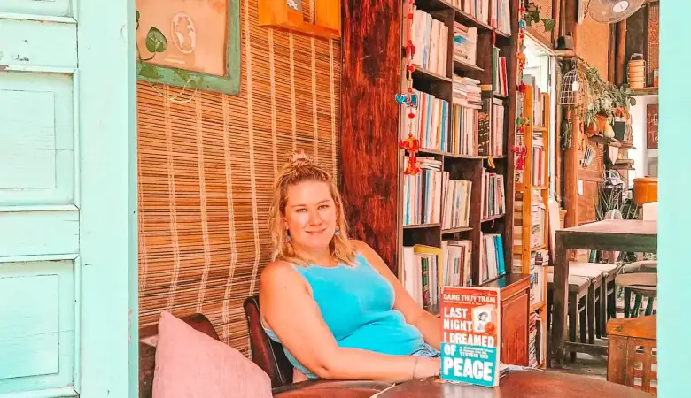A woman in a blue top relaxes by a turquoise window in a book café, reading Last Night I Dreamed of Peace by Dang Thuy Tram. Bookshelves and fan-cooled walls give the space a homey, thoughtful atmosphere.