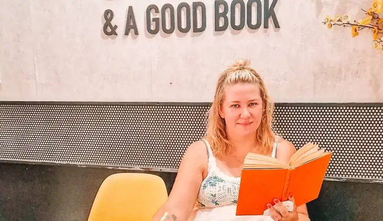Em relaxes in a modern café with a coffee in one hand and an orange book in the other, titled 'The Refugees'. She sits under a wall sign that reads "Happiness is a cup of coffee & a good book."