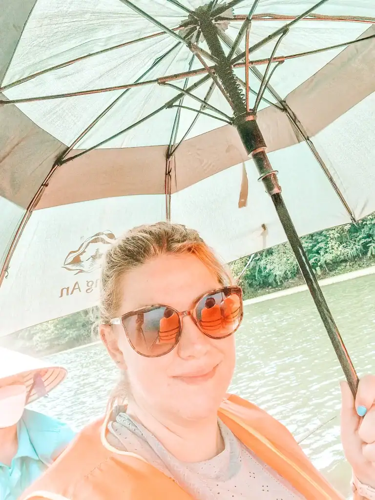 Close-up of a woman holding a large umbrella for shade while wearing sunglasses and a life vest on a boat ride in Ninh Binh. Another rower in a conical hat can be seen in the background.