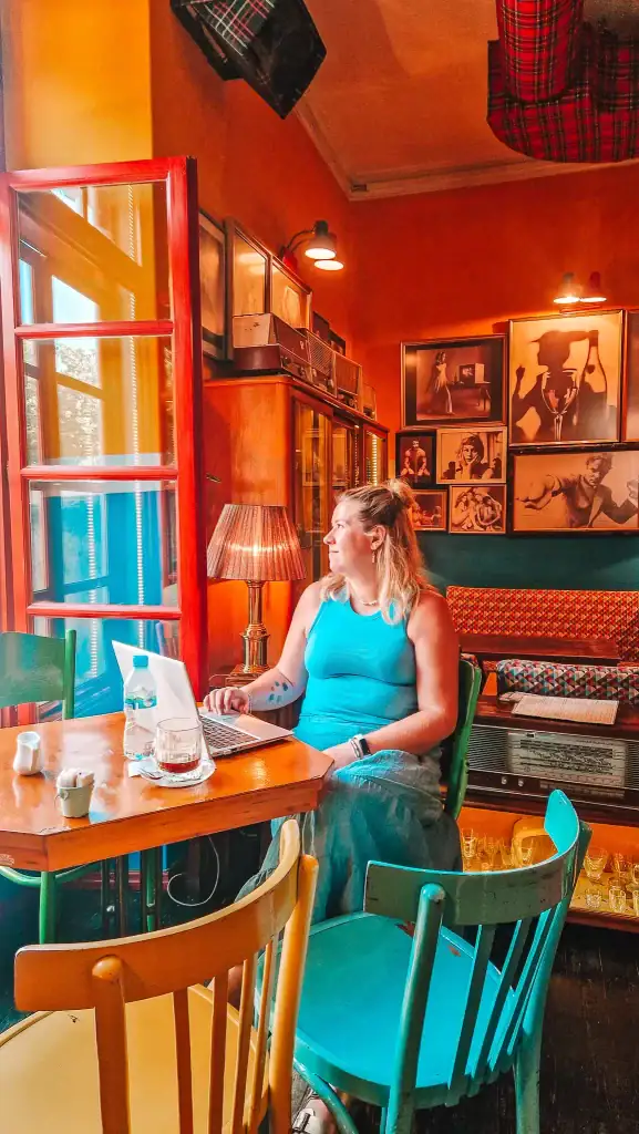 Em, dressed in turquoise, works on her laptop in the vibrant and vintage-inspired interior of Radio Bar in Tirana. Surrounded by colorful furniture, retro decor, and framed black-and-white photography, this café feels like a creative time capsule.
