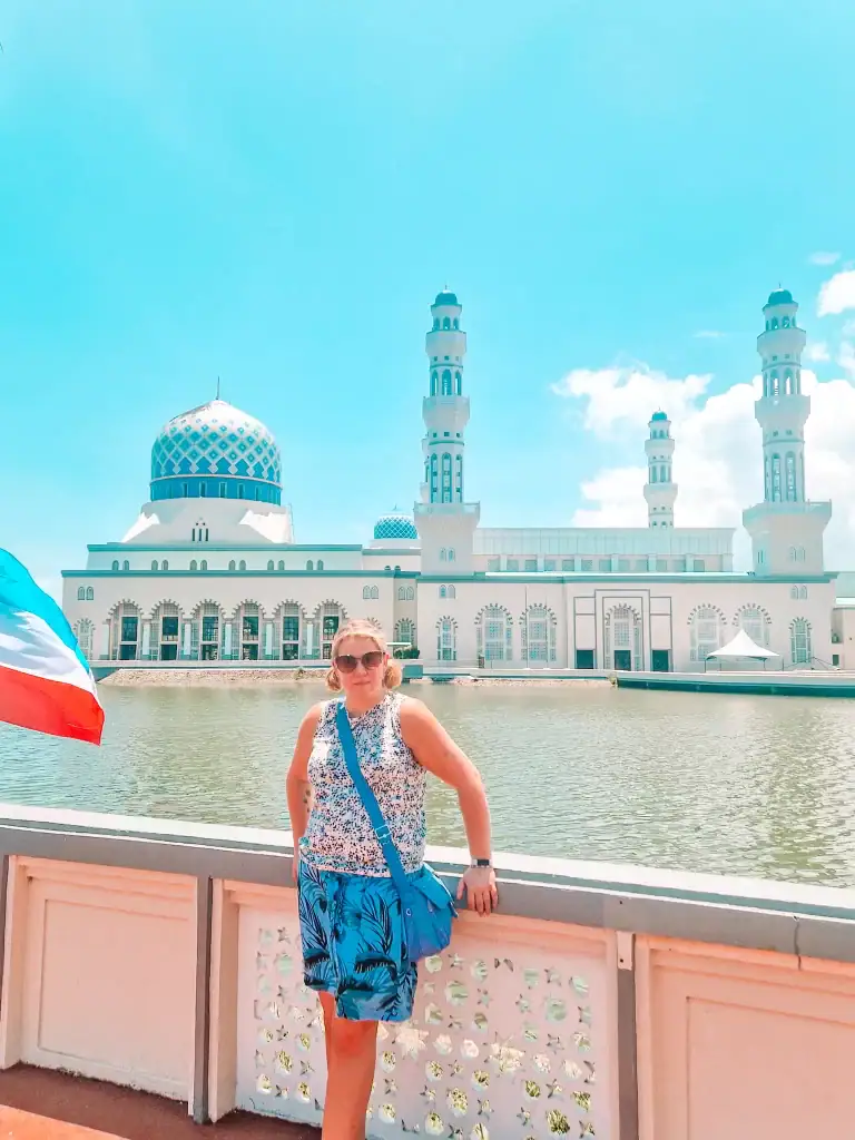 Em poses by the railing in front of the majestic blue-domed Kota Kinabalu City Mosque, reflecting in the calm waters under a clear turquoise sky.