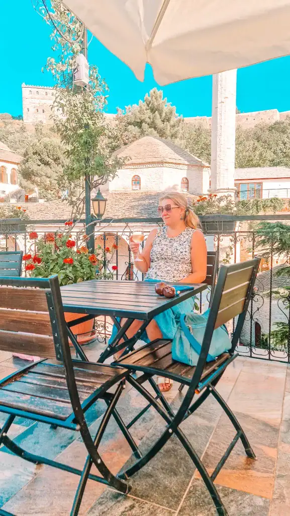 Em enjoys a glass of wine at a shaded café terrace in Gjirokaster’s old town, with Ottoman architecture and the castle in the background.