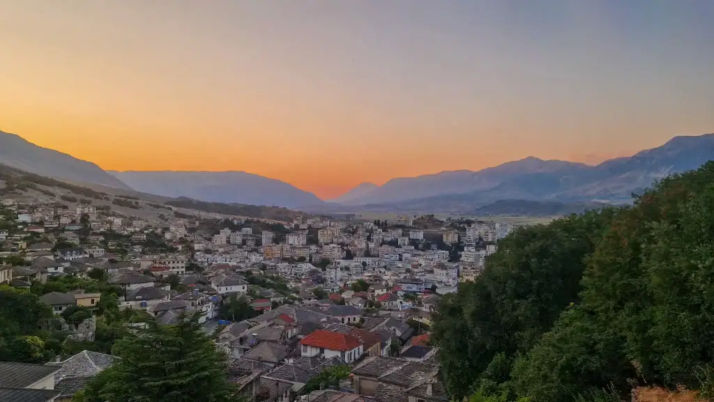 A panoramic view of Gjirokaster at sunset, with the city nestled in a wide valley between mountain ranges under a glowing orange sky.