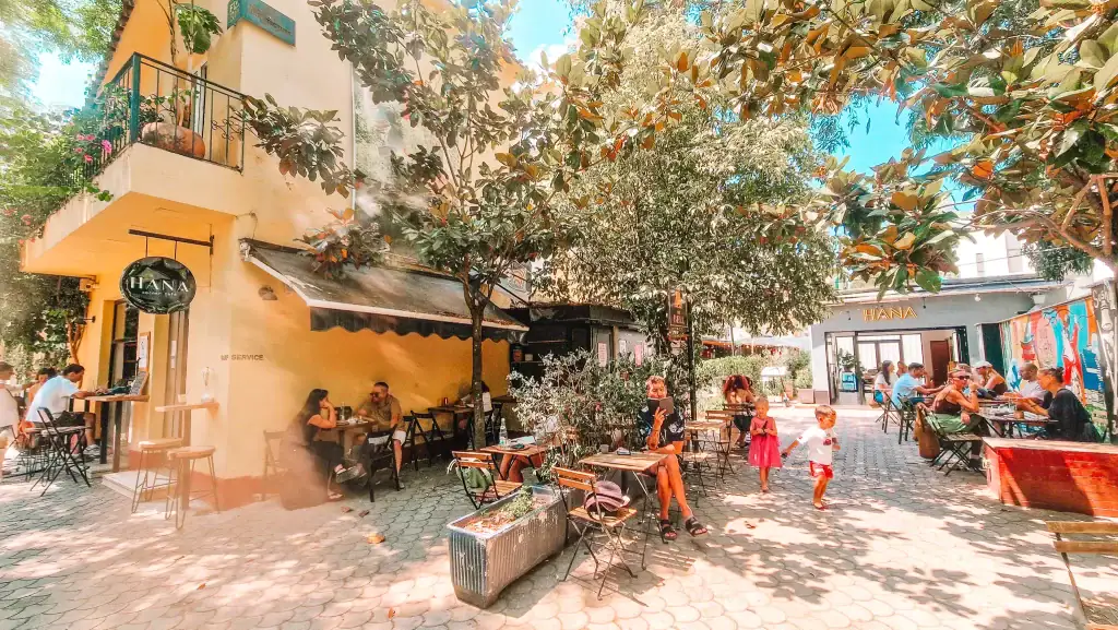 Bustling outdoor scene at Hana Café in Tirana, with diners chatting under leafy trees and kids playing in the sun-dappled courtyard. The lively yet cozy setting places it high on the list of best cafes in Tirana.